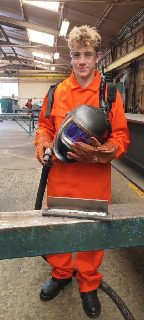 A man in an orange suit is holding a welding helmet in a warehouse.