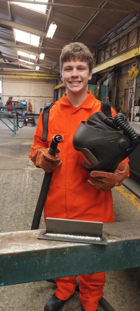 A young man in an orange jumpsuit is holding a welding machine in a factory.