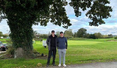 Two men are standing under a tree in a grassy field.