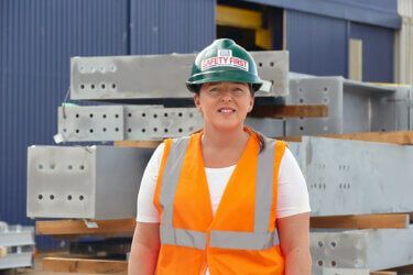 A woman wearing a hard hat and an orange vest is standing in front of a pile of metal.