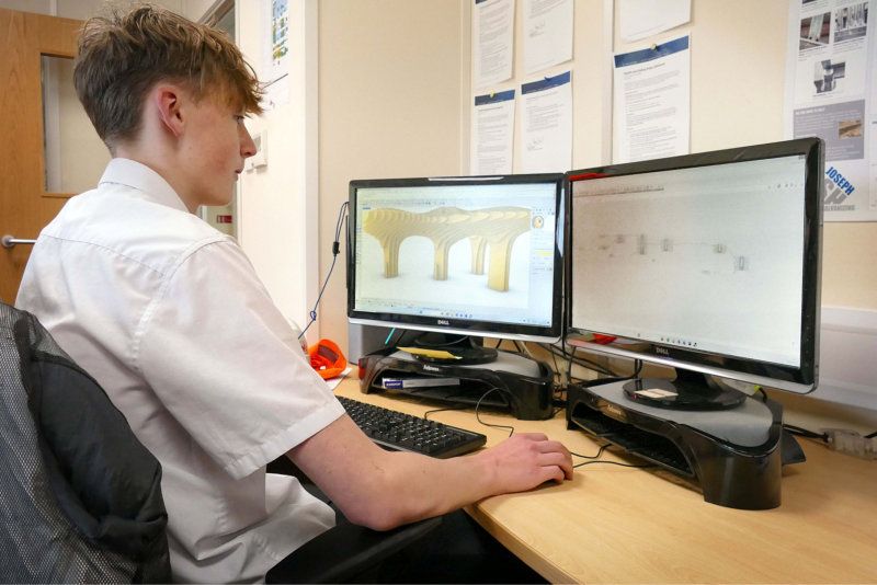 A young man is sitting at a desk in front of two computer monitors.