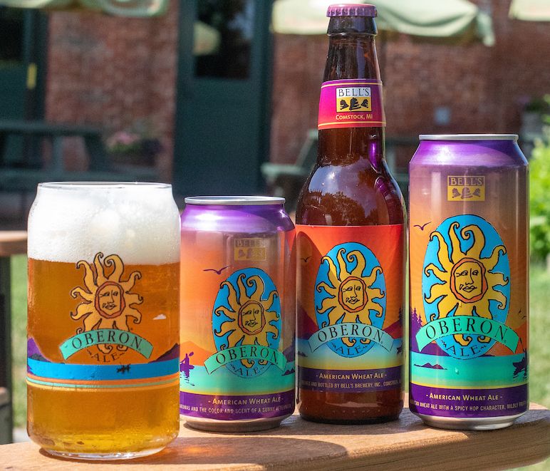 Oberon beer bottles, cans, and glass with foamy beer, set outdoors. Sunny label with a face.