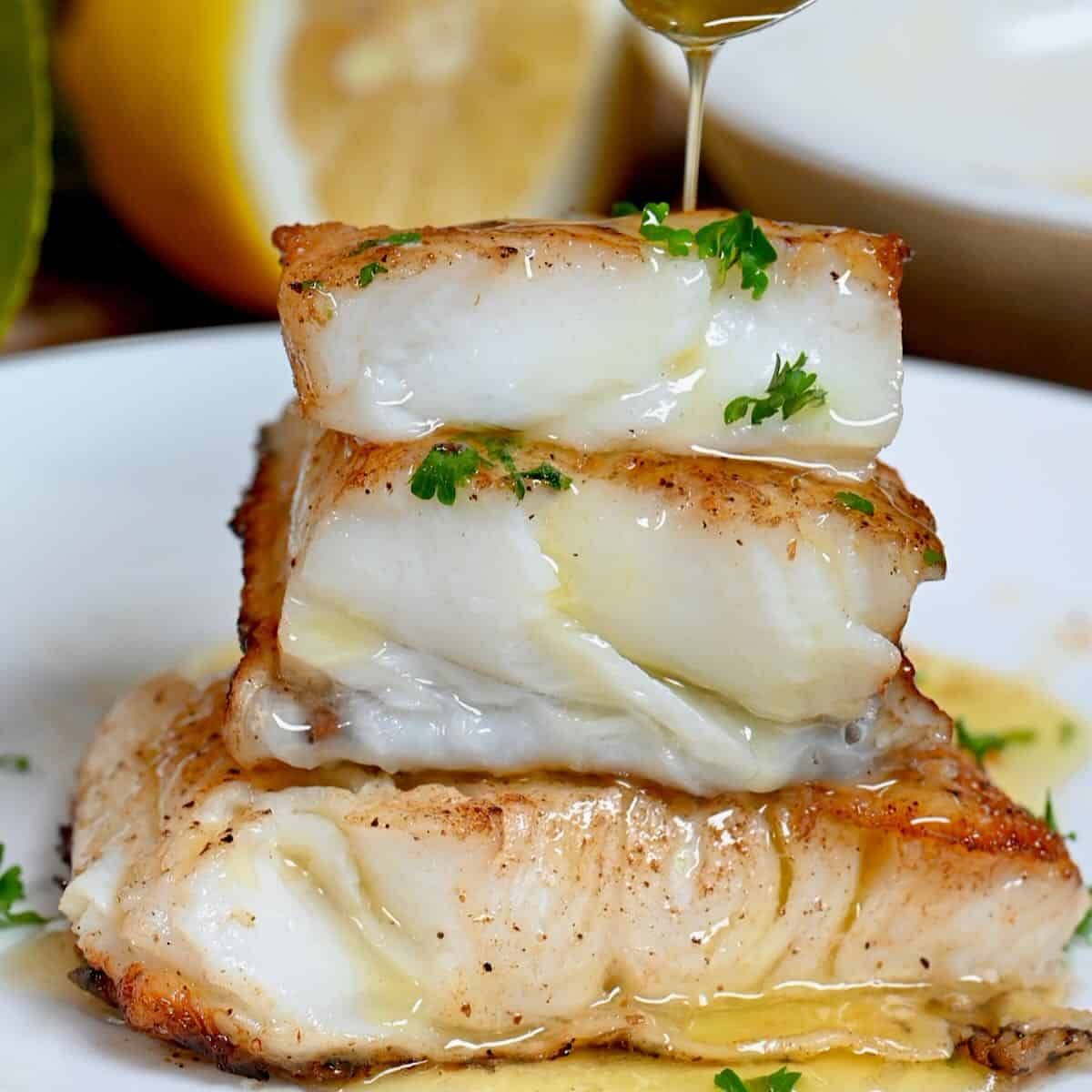 Pan-seared cod stacked on a plate with sauce being drizzled on top, garnished with parsley and lemon.