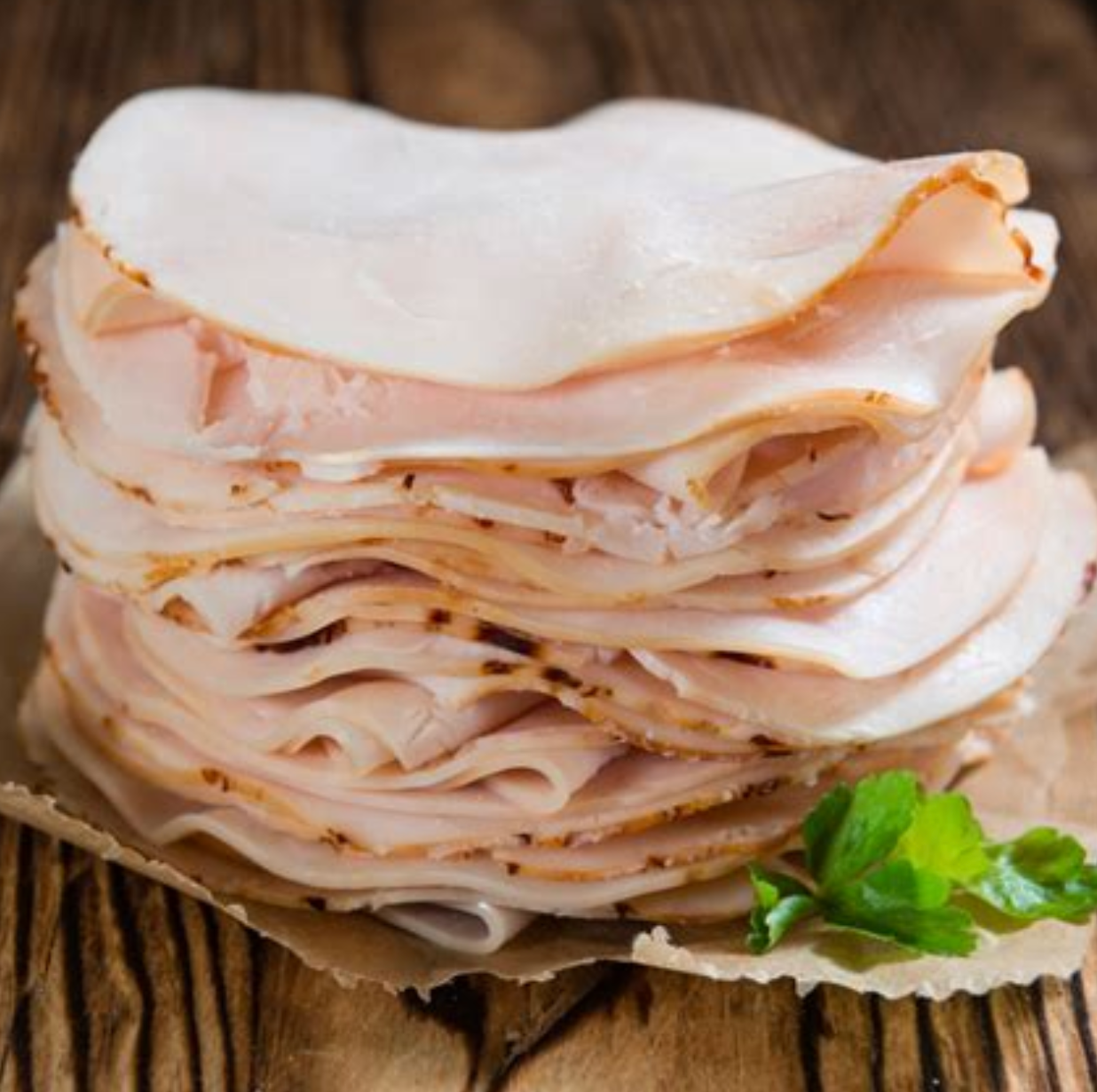 Stack of sliced turkey on parchment paper, wood table, some parsley.