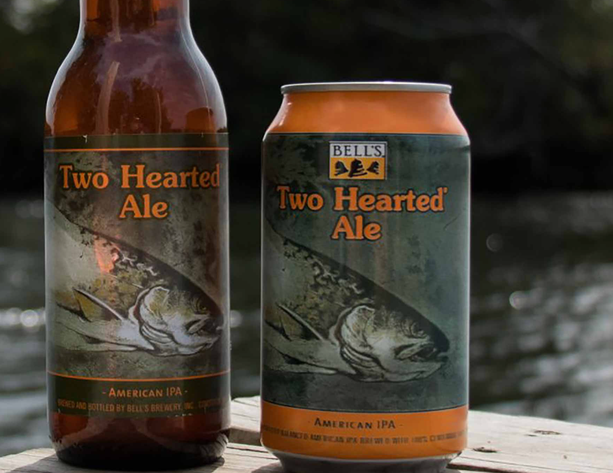 Two Bell's Two Hearted Ale bottles & can with fish graphic, amber color, set near water.