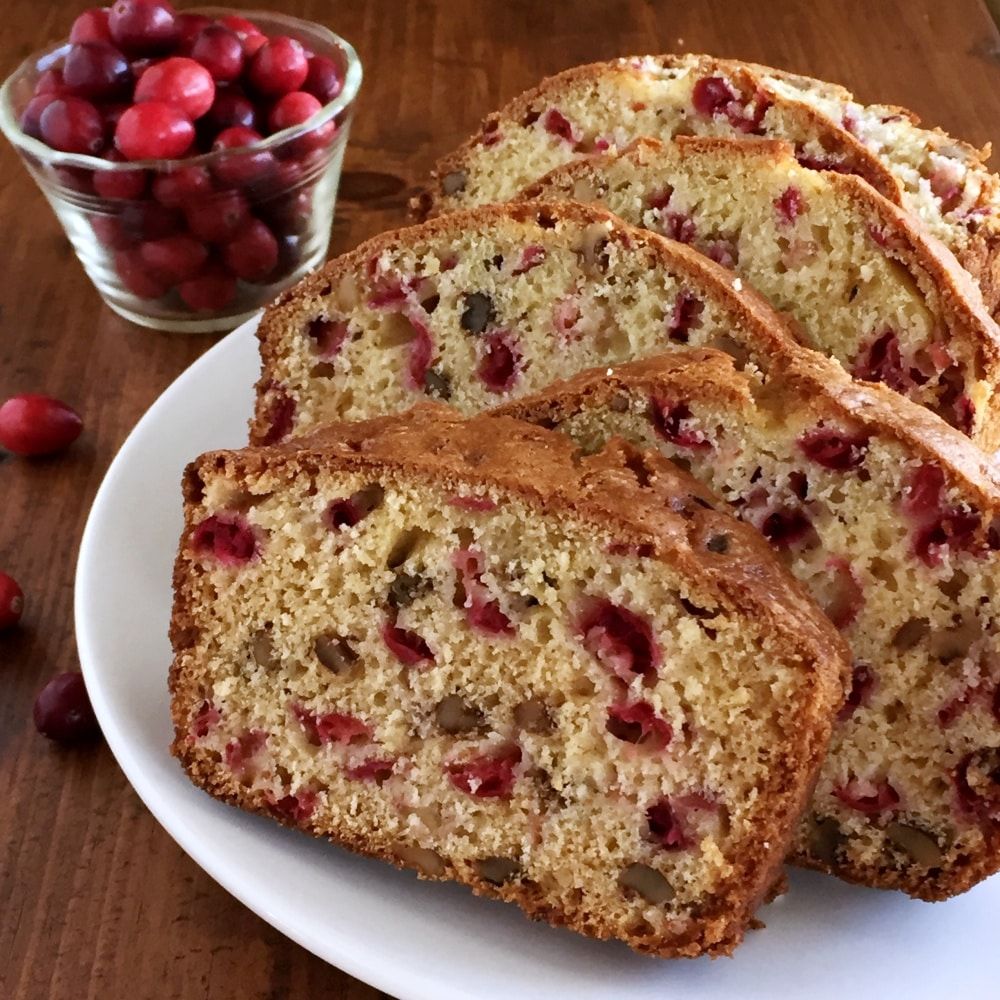 Sliced cranberry walnut bread on a white plate, with a bowl of cranberries on a wooden surface.
