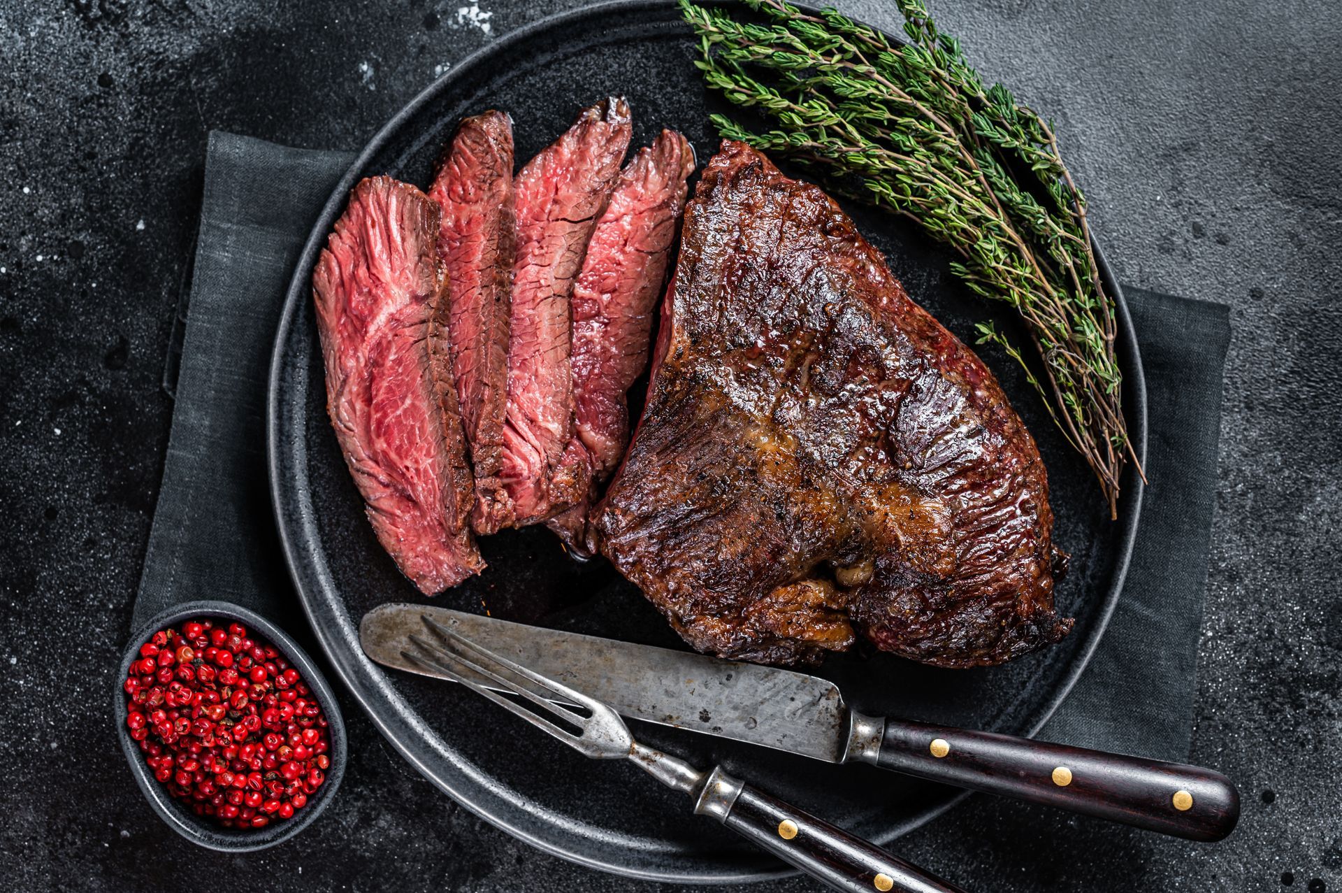 Sliced, seared tri-tip steak on a black plate with red peppercorns and thyme.