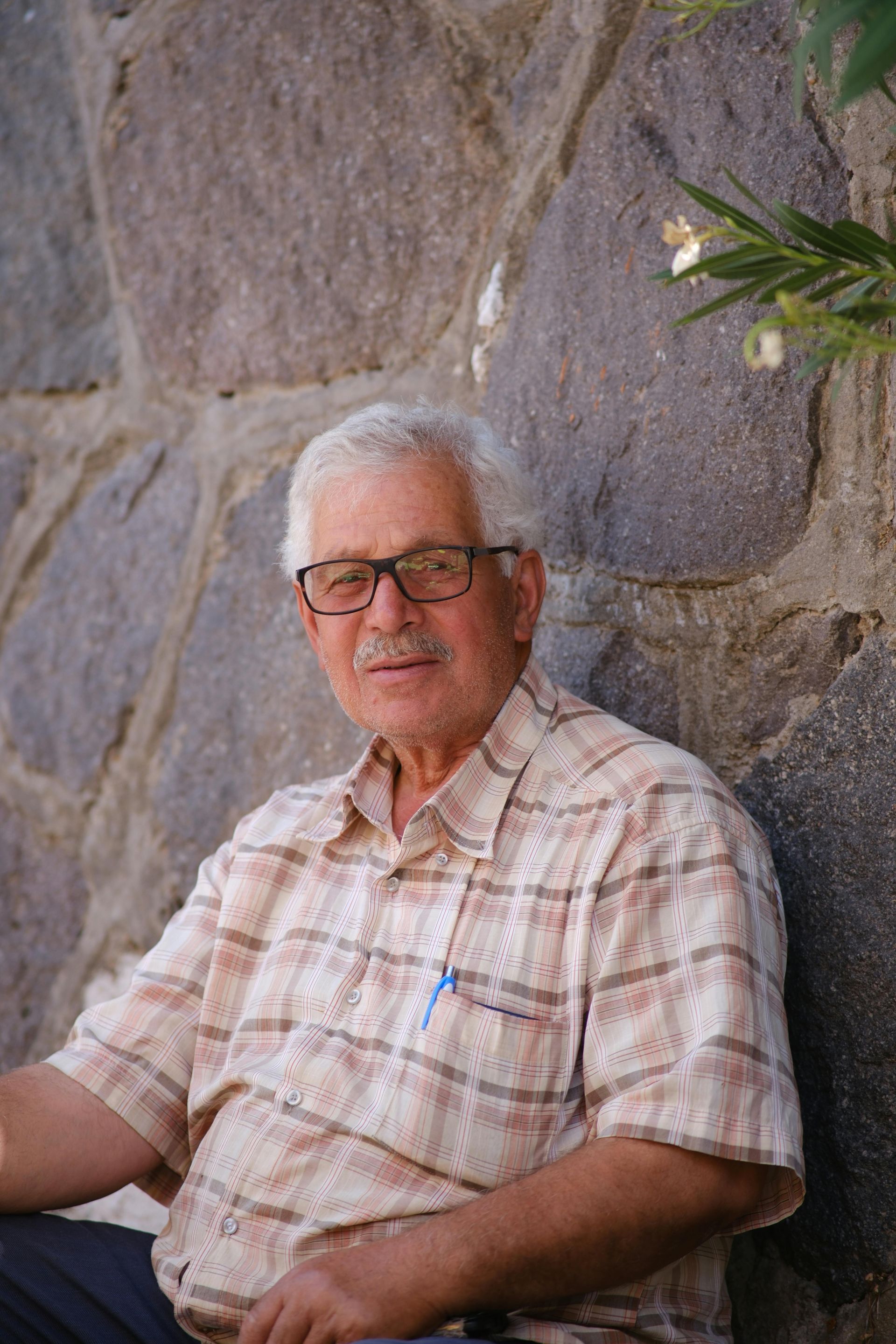 Man with glasses and mustache in patterned shirt sits in front of a stone wall.