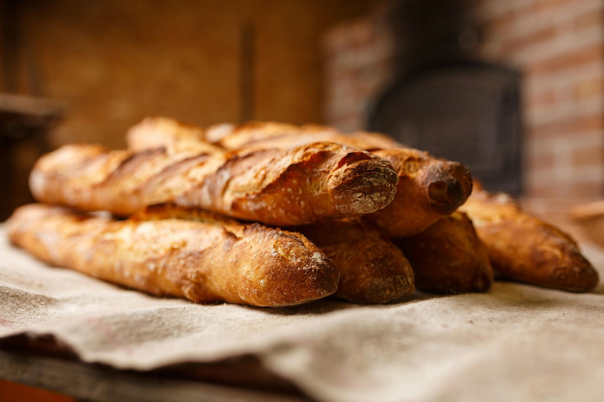 Pile of golden baguettes on a linen cloth, set in front of a brick oven.