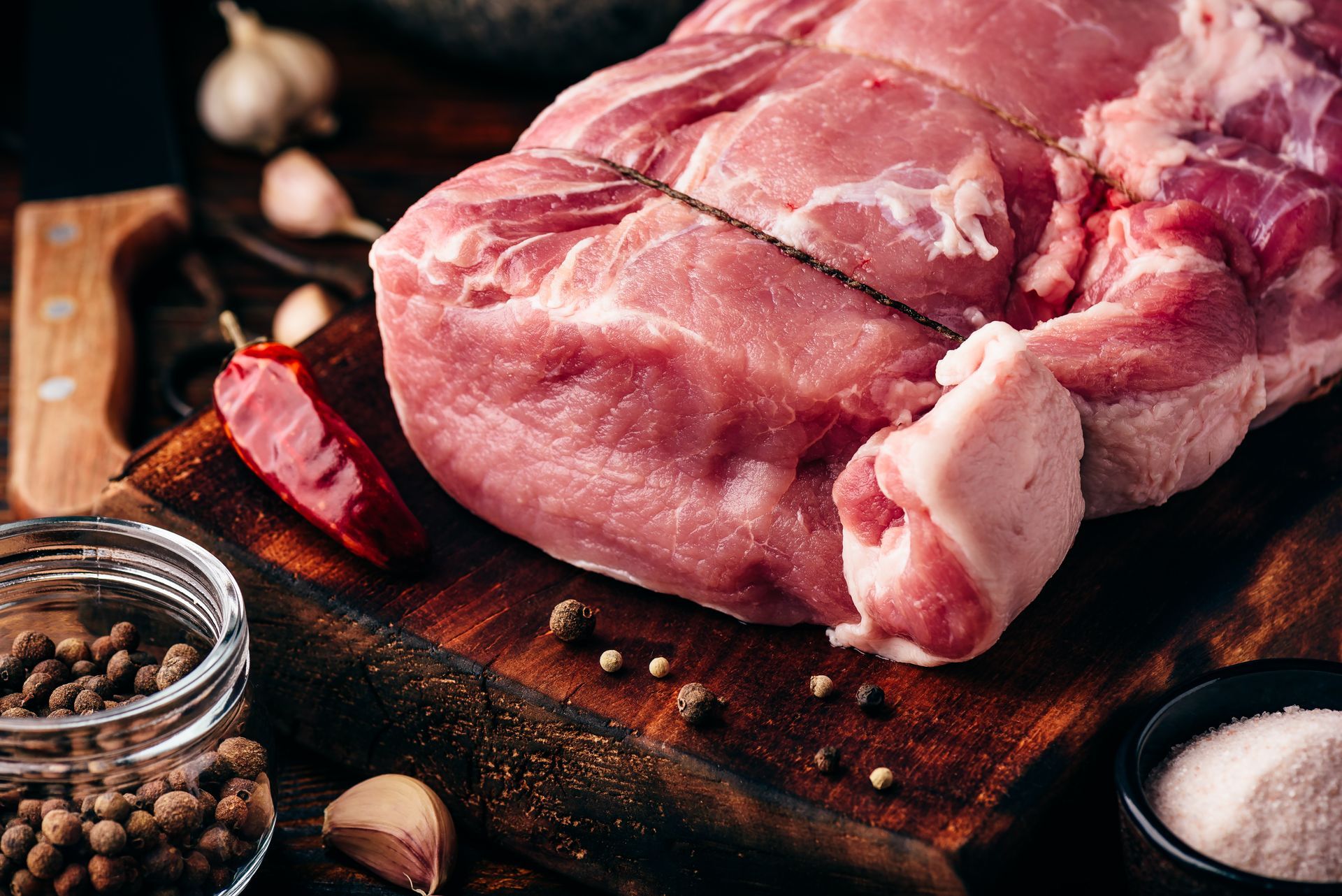 Raw pork roast tied with twine, on a wooden cutting board with spices: peppercorns, garlic, salt, chili pepper.