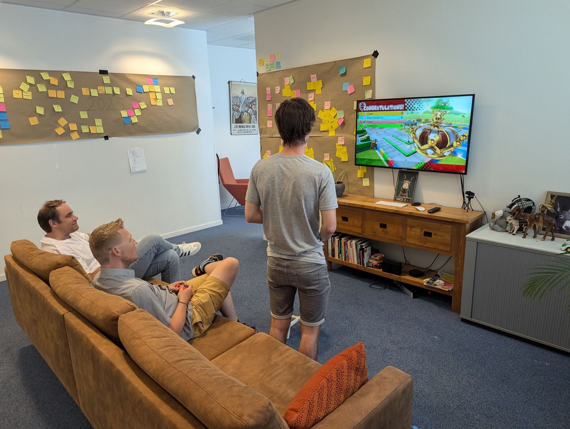 Drie mensen zitten in een woonkamer en kijken naar een kleurrijk videospel op de televisie.
