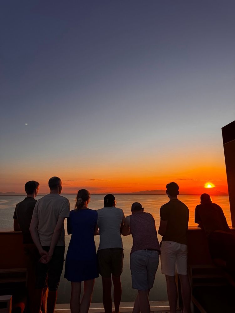 Zes mensen staan op een balkon en kijken naar de zonsondergang.