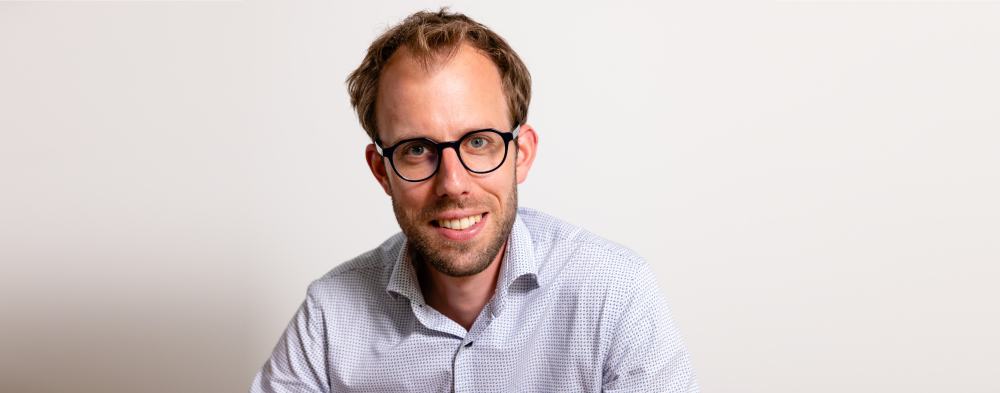 Portret van een lachende man met bril in een lichtblauw shirt tegen een witte achtergrond.