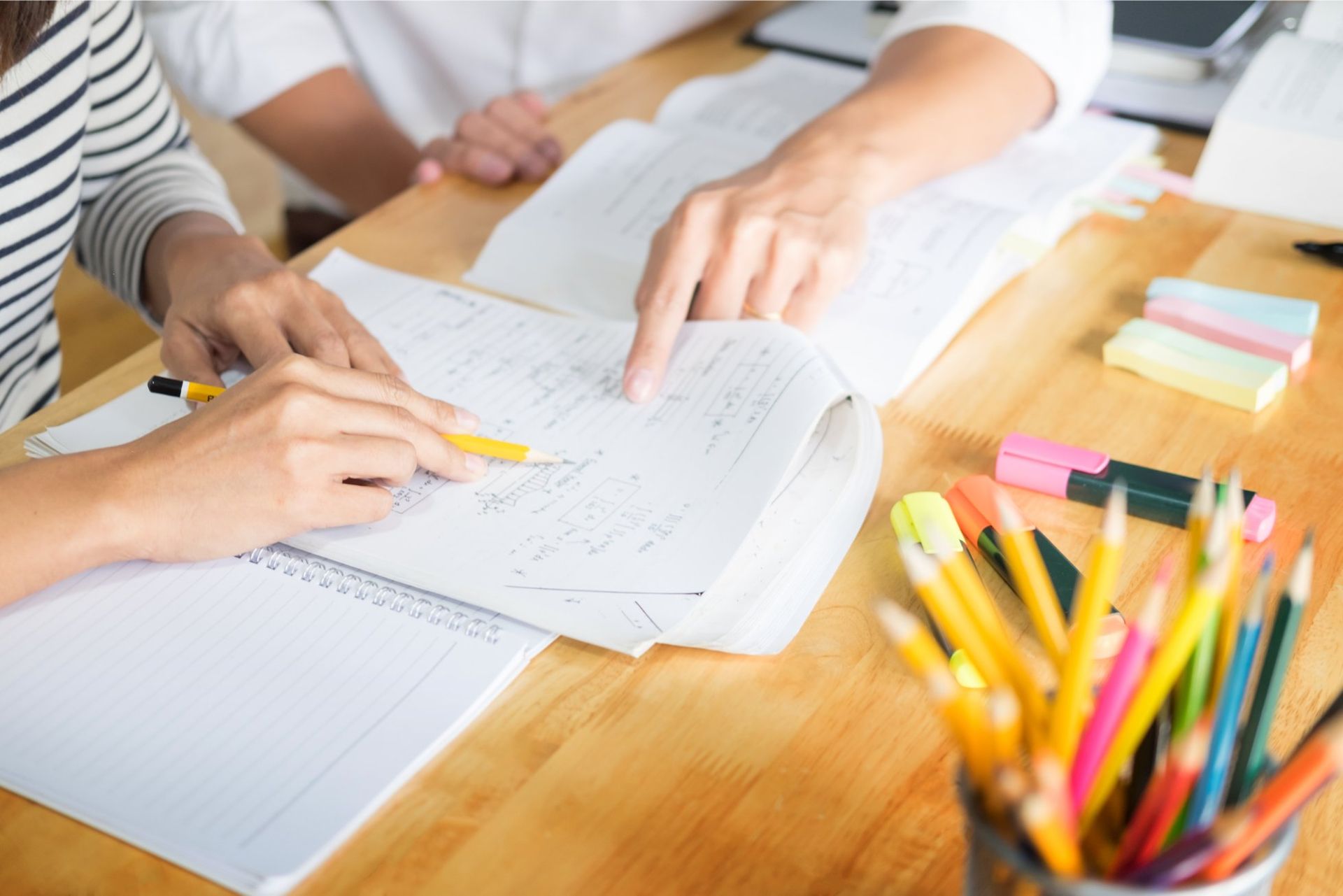 Two people pointing at notes and papers on a wooden table, studying. Colored pencils in a cup.