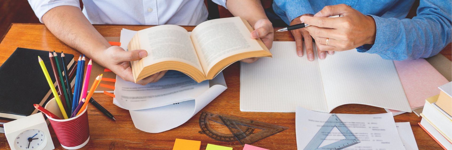 Two people studying together, one holding a book open, the other with a pen and paper.