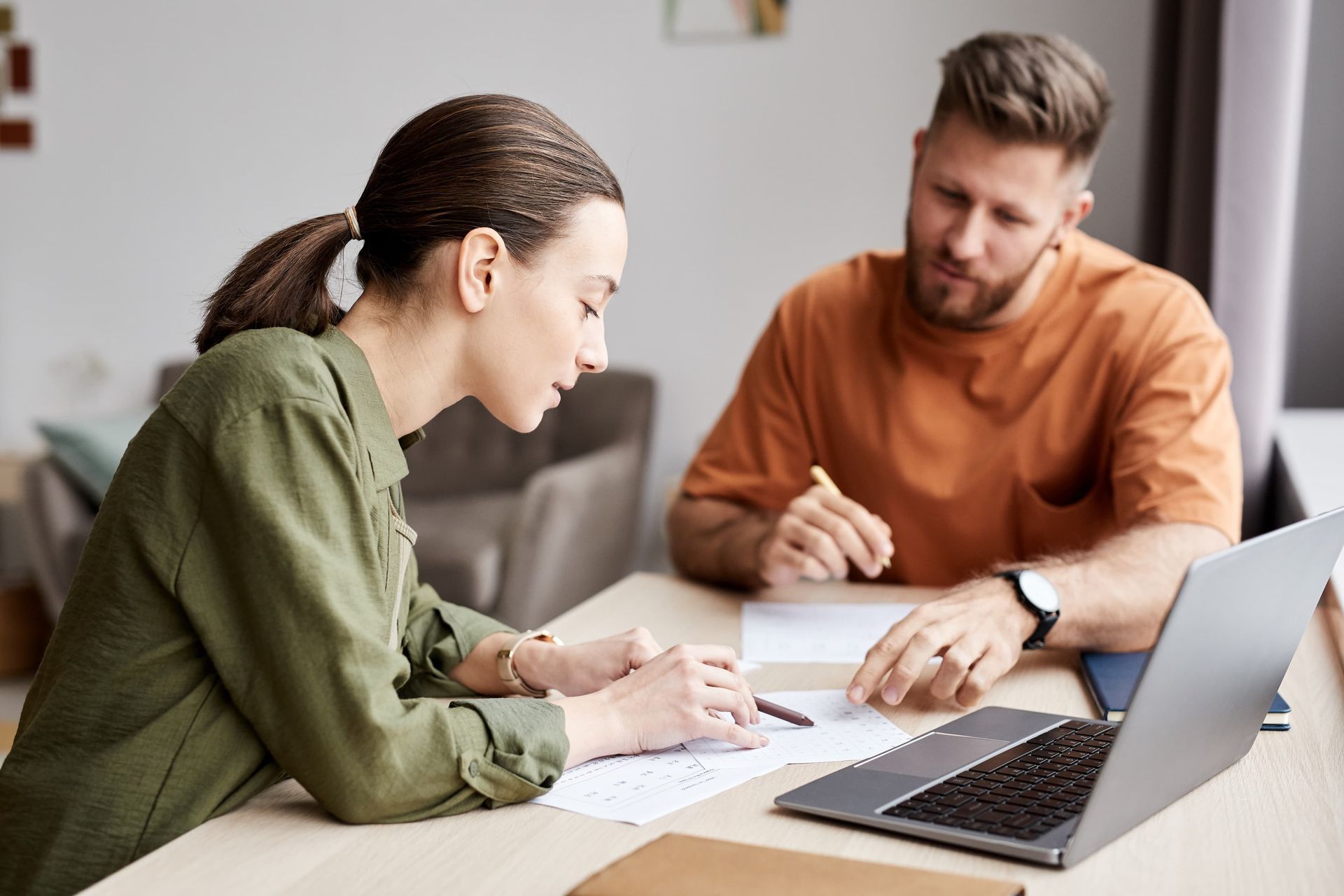 Woman in green shirt and man in orange shirt reviewing papers at a table with a laptop.