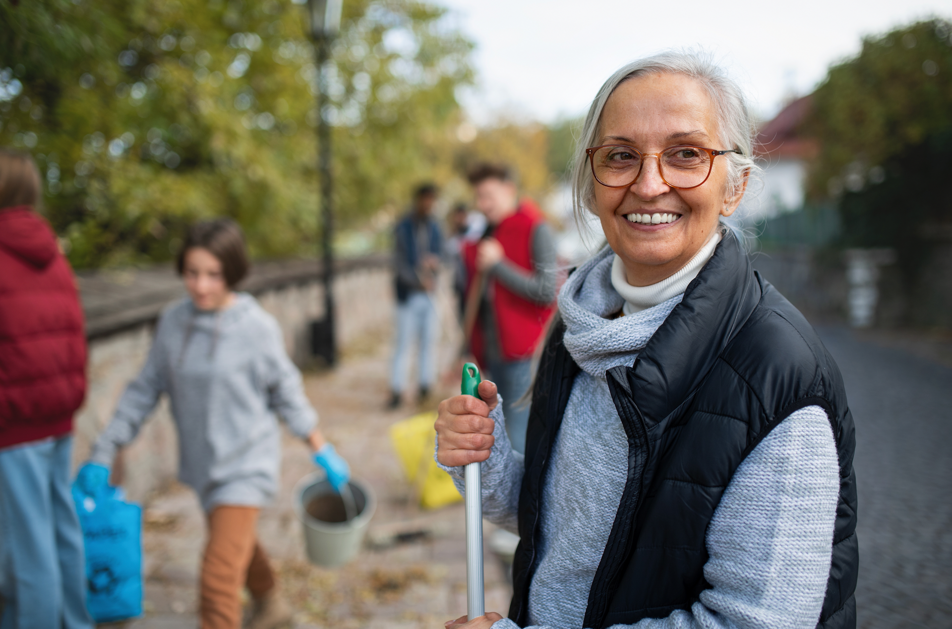 An elderly woman is holding a broom and smiling while standing in front of a group of people.