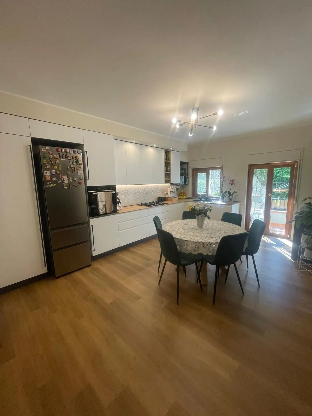 Luminosa cucina moderna con zona pranzo, pavimento in legno, sedie nere, mobili bianchi e porte finestre che danno sul patio.