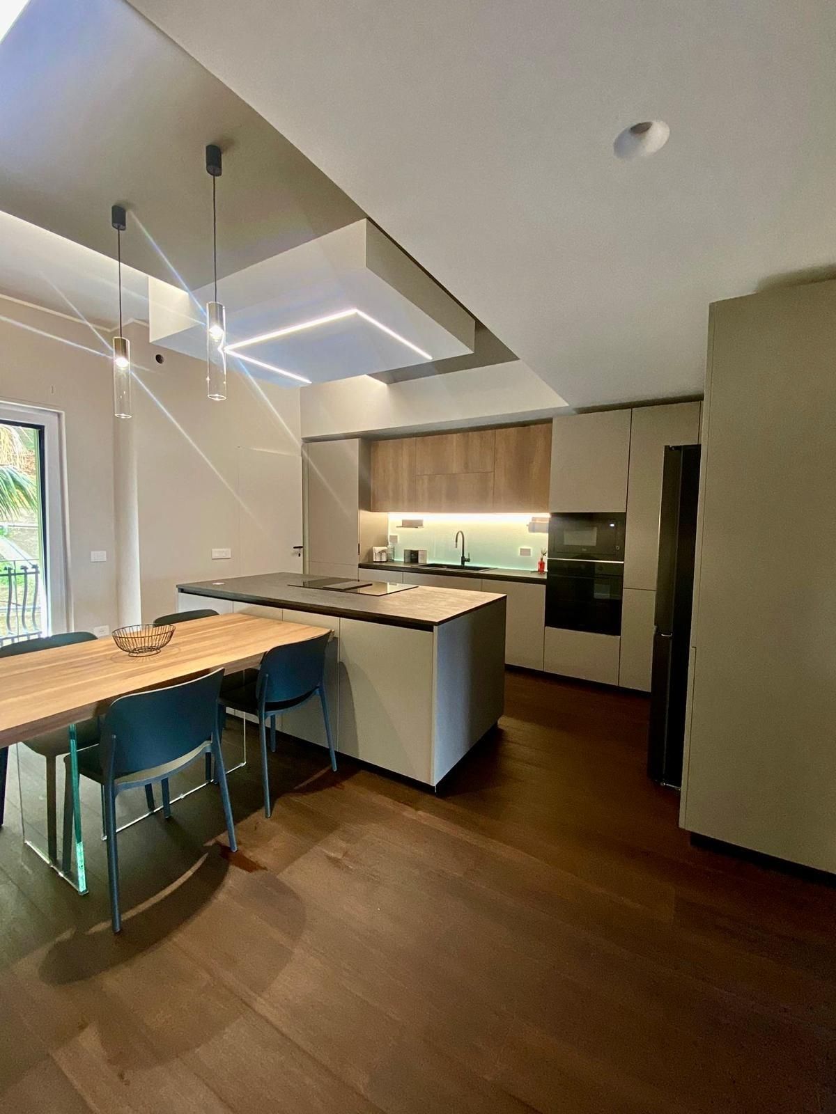 Cucina moderna con isola, tavolo da pranzo in legno, lampade a sospensione e pavimento in legno scuro.