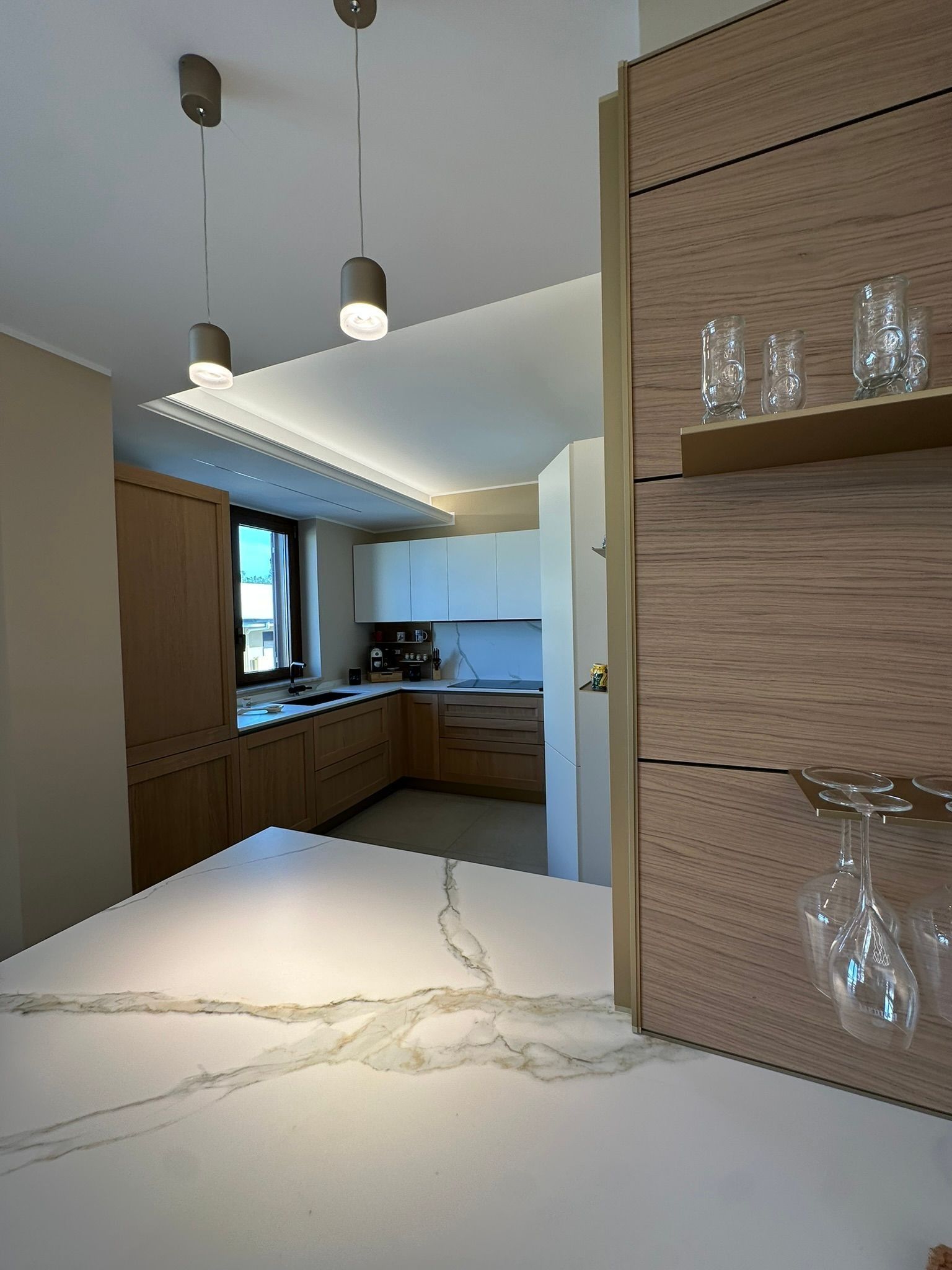Cucina moderna con mobili in legno, isola in marmo bianco, lampade a sospensione e mensole a vista con bicchieri.
