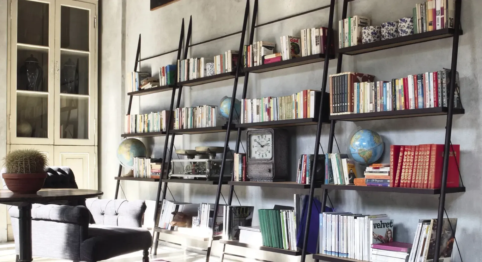 Luminoso soggiorno con librerie in metallo nero, libri, oggetti decorativi, un mappamondo e un tavolino vicino alla finestra.