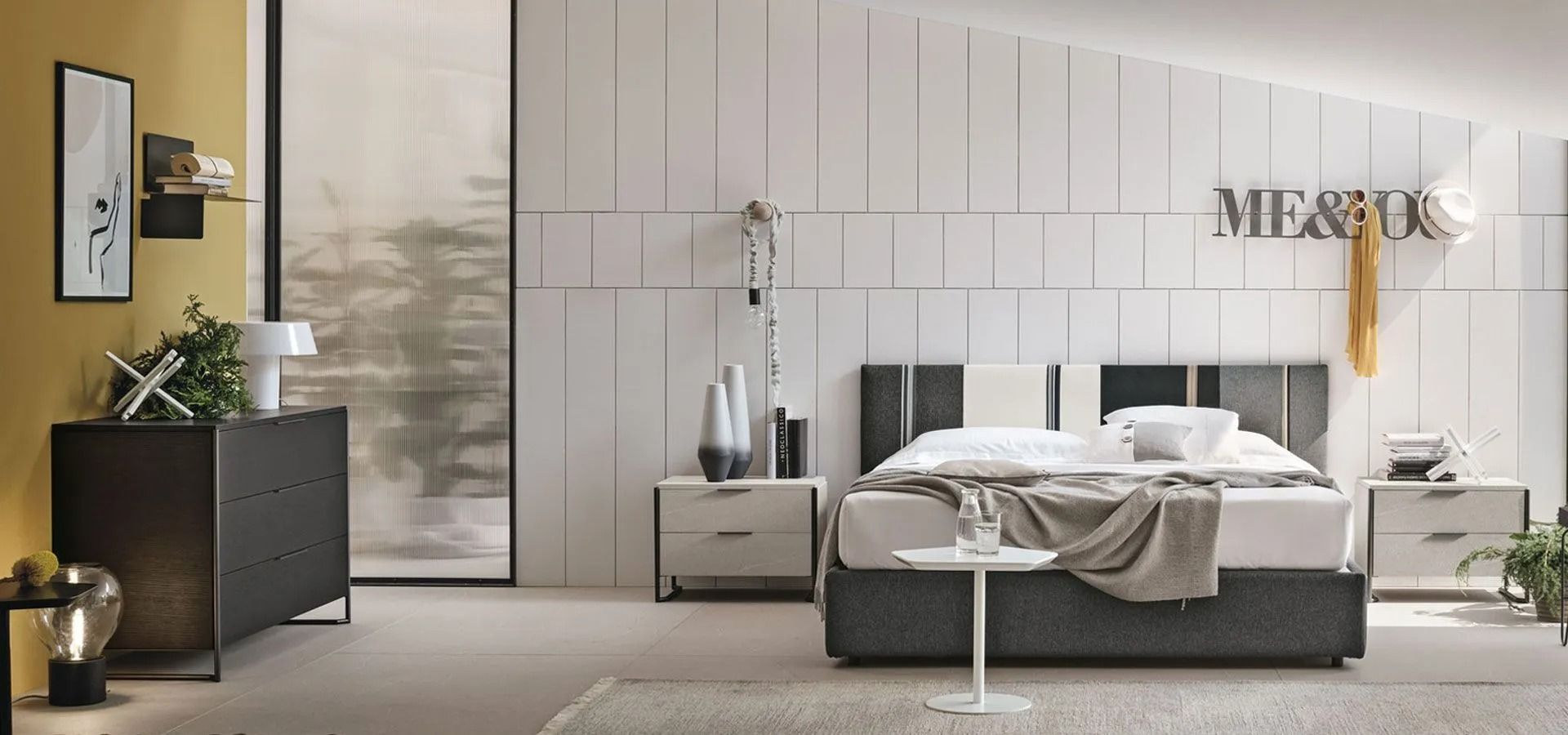 Camera da letto moderna con letto grigio, parete rivestita in pannelli bianchi, lampada dorata e comò nero con pianta.