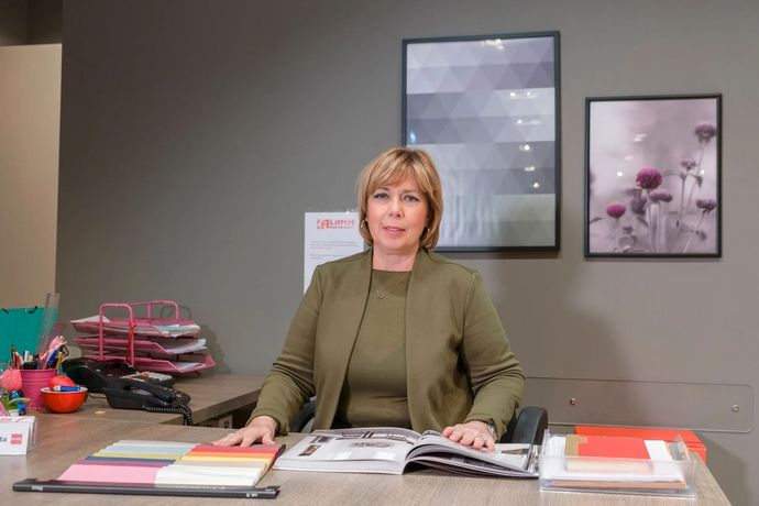 Donna seduta a una scrivania in un ufficio, con libri e documenti aperti, rivolta verso la telecamera.