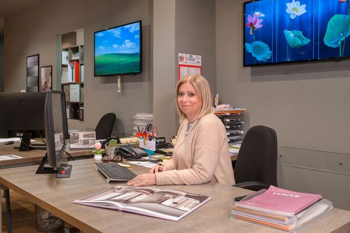 Donna seduta a una scrivania in ufficio, guarda la telecamera con monitor di computer e documenti nelle vicinanze.