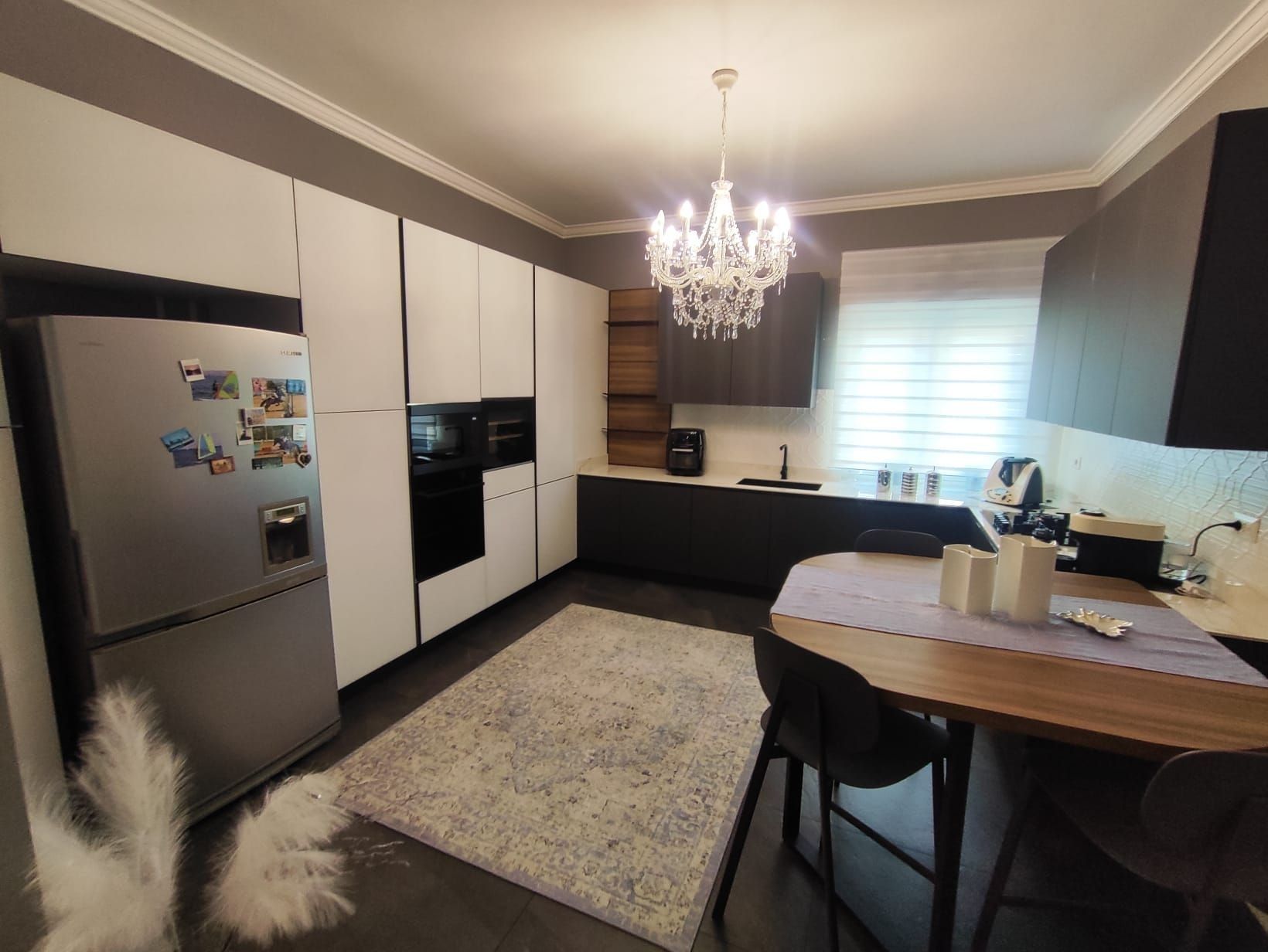Cucina moderna con frigorifero in acciaio inox, mobili in bianco e nero, lampadario, finestra e tavolo da pranzo in legno.