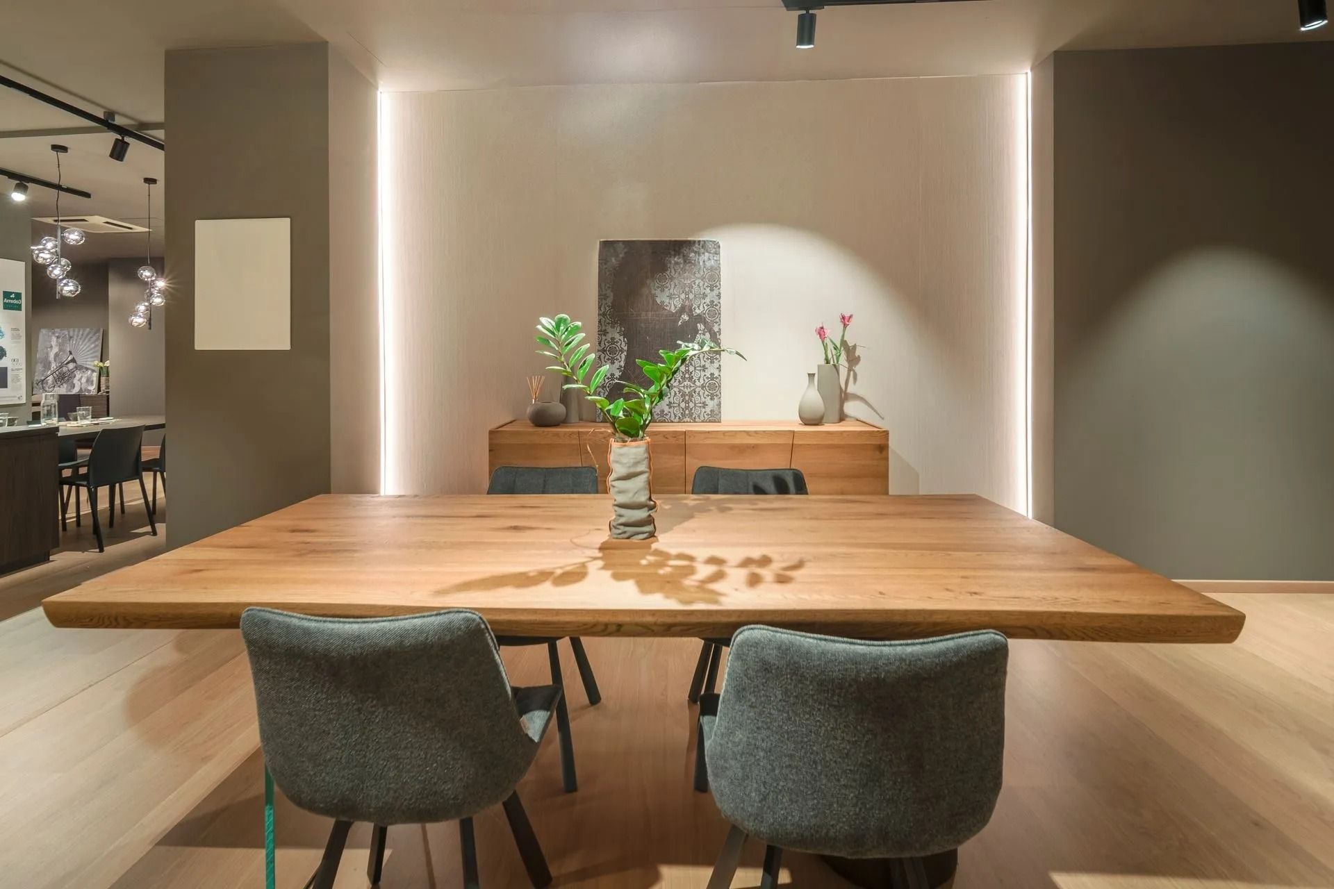 Sala da pranzo moderna con tavolo in legno, due sedie grigie e una piccola composizione di piante sotto luci calde.