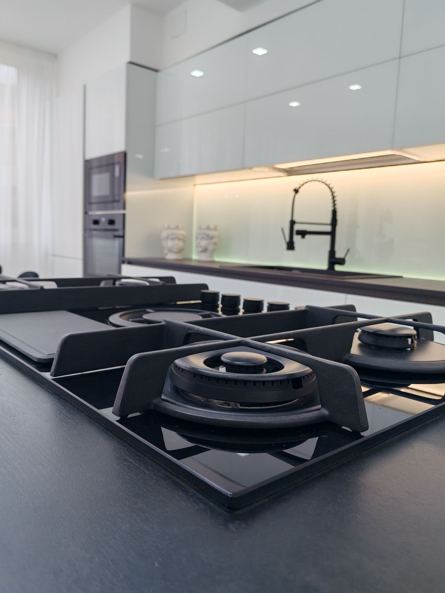 Cucina moderna bianca con piano cottura a gas nero e forno a incasso, mobili lucidi e lavello sotto luci sottopensile.