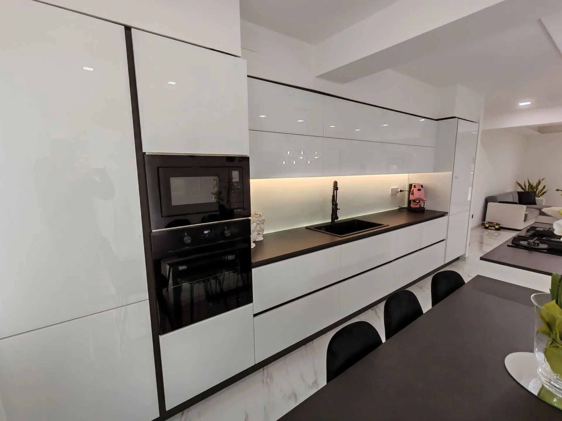 Cucina moderna bianca con forno a incasso nero, lungo bancone, lavello e sgabelli a isola.