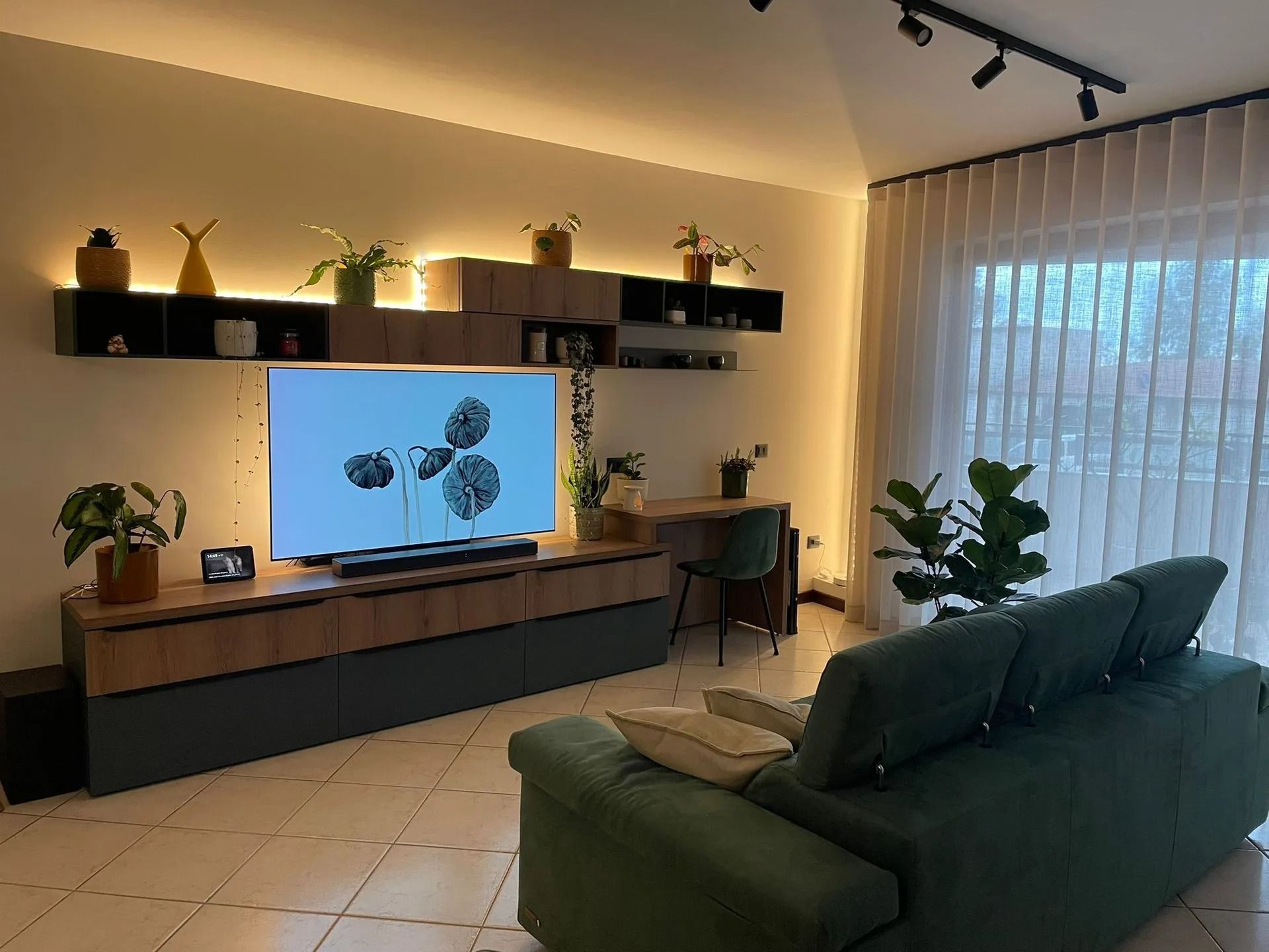 Soggiorno moderno con divano verde, TV a parete, illuminazione calda, mensole e scrivania vicino a una finestra.