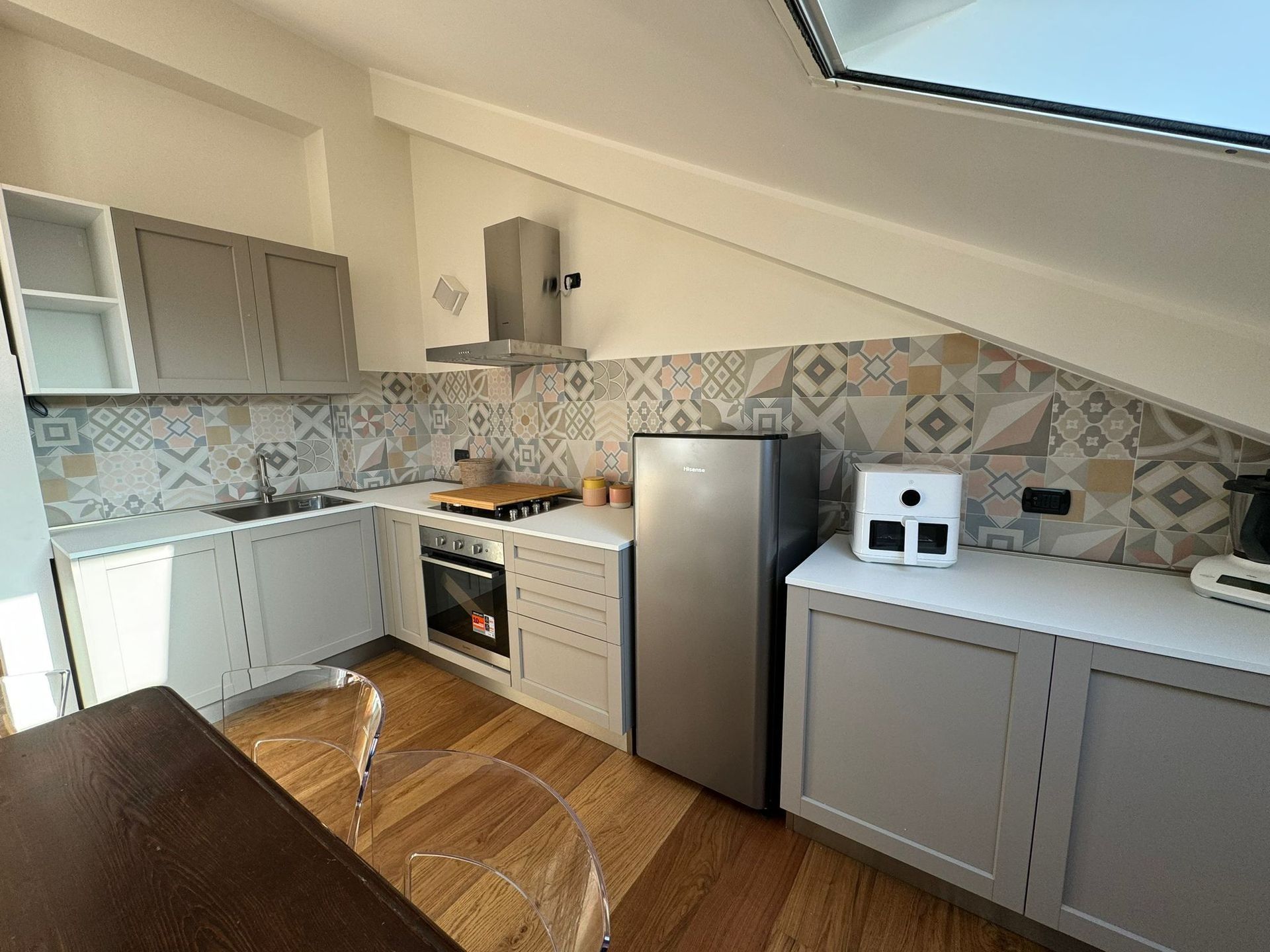 Piccola cucina moderna con mobili grigi, elettrodomestici bianchi, paraschizzi decorato e lucernario.