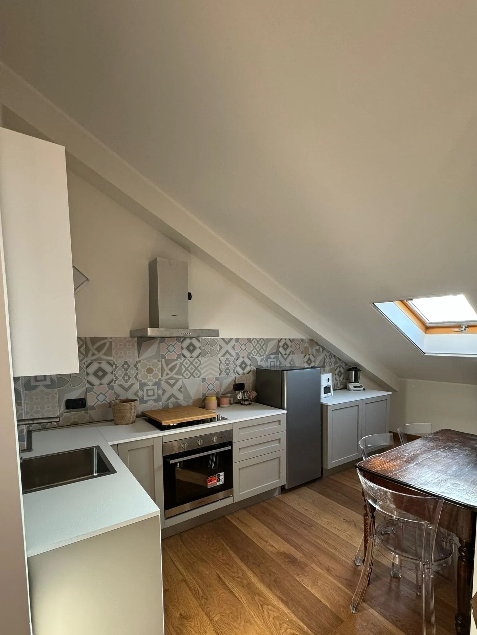 Piccola cucina mansardata con mobili bianchi, pavimento in legno, paraschizzi a mosaico e lucernario.