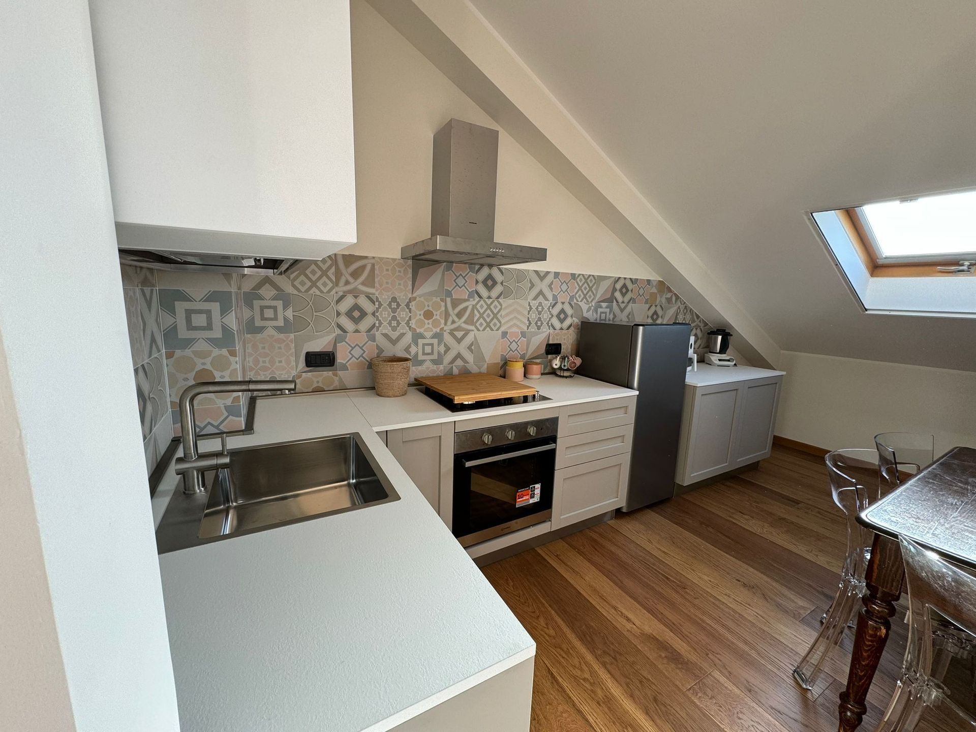Cucina compatta in mansarda con mobili bianchi, paraschizzi decorato, pavimento in legno e lucernario.