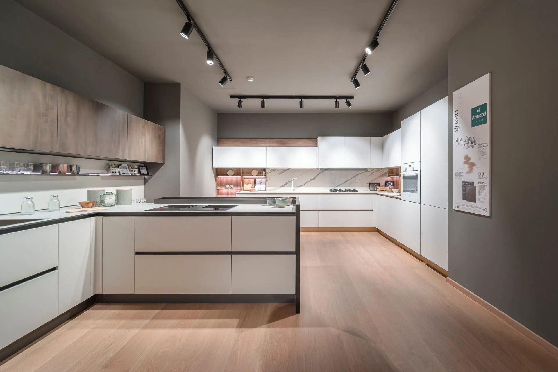 Showroom di cucine moderne bianche con isola, mobili, pavimento in legno e faretti a binario.