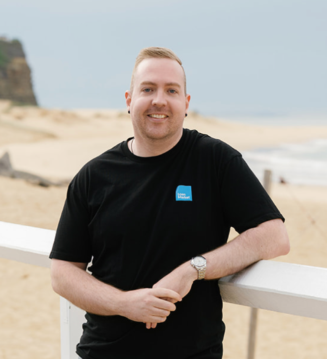 Man smiling, leaning on a white railing at a beach. Black t-shirt, watch, sandy beach in background.