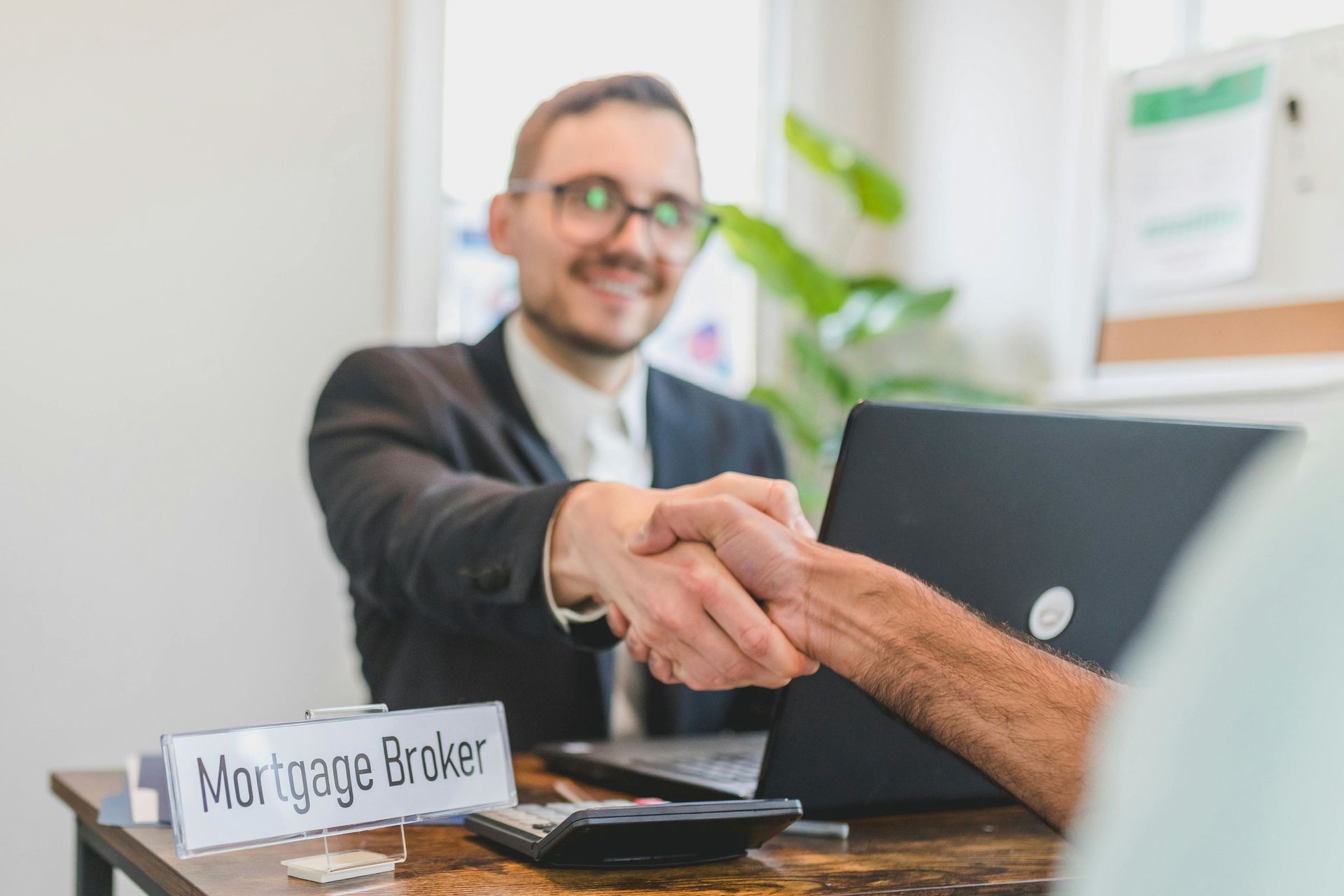 Mortgage broker shaking hands with a client at a desk, laptop and nameplate visible