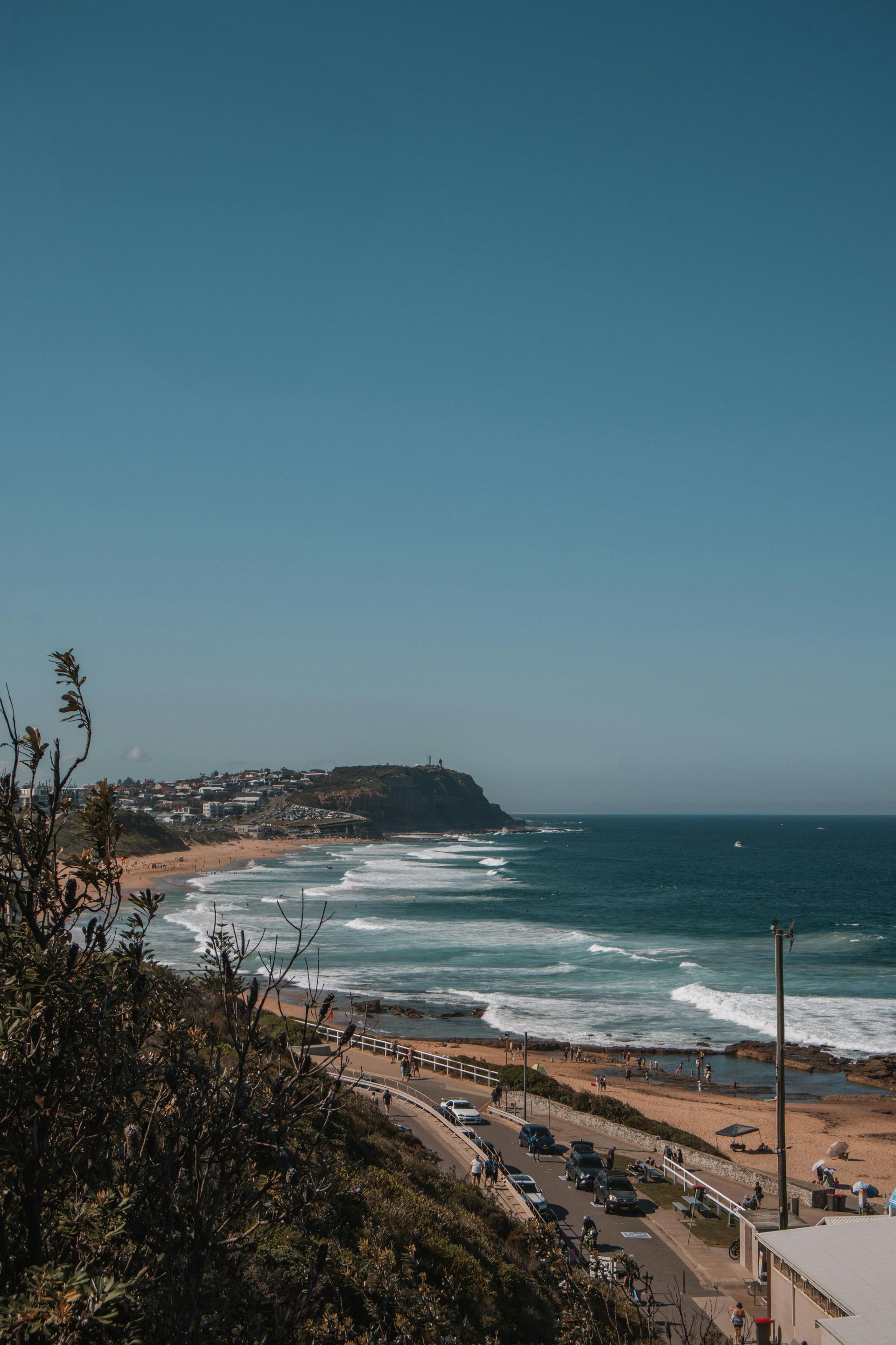 Coastal road and beach with waves under a clear blue sky, viewed from a hillside.