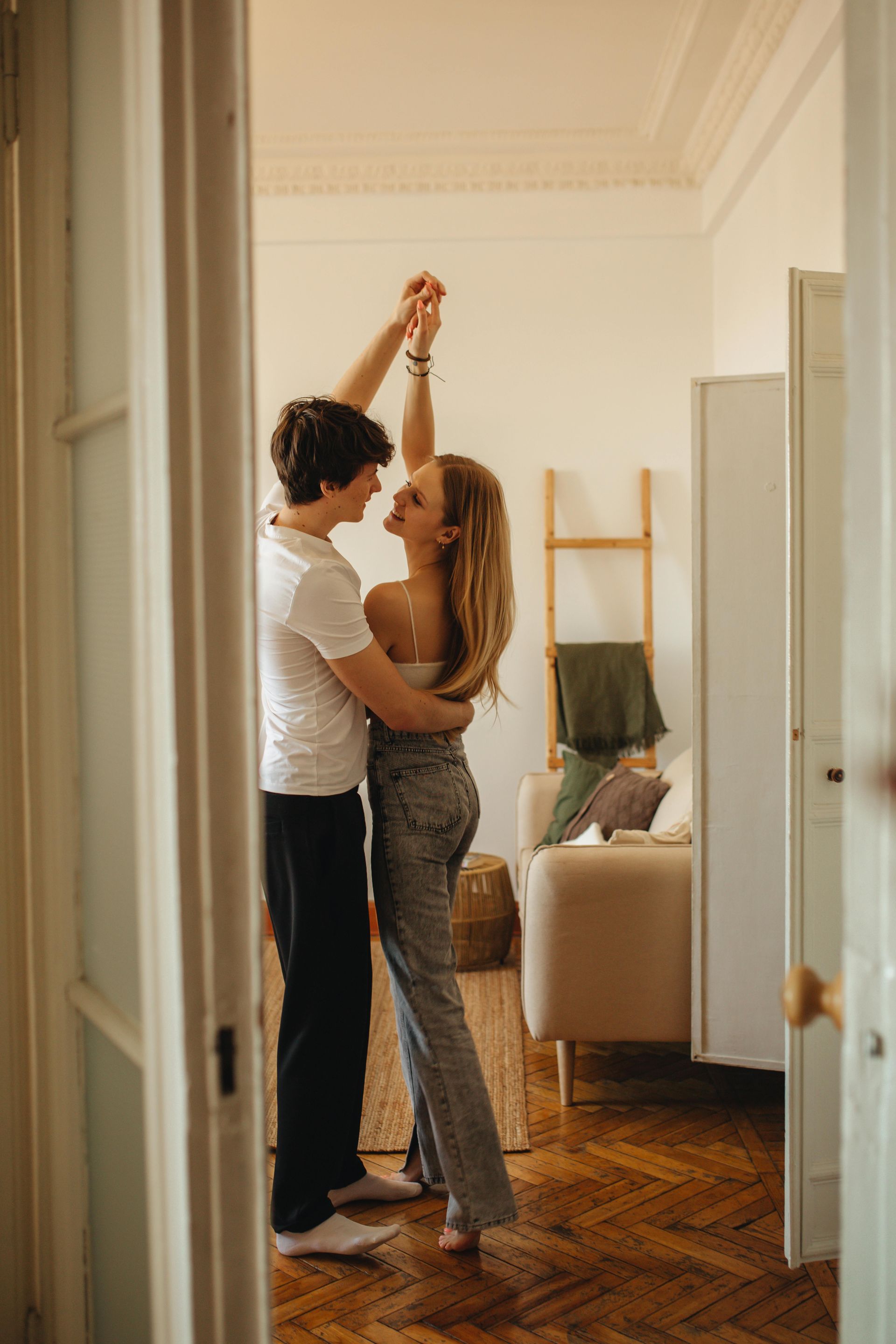 Two people dance in a bright living room, one leading with raised hands near a sofa and ladder shelf.