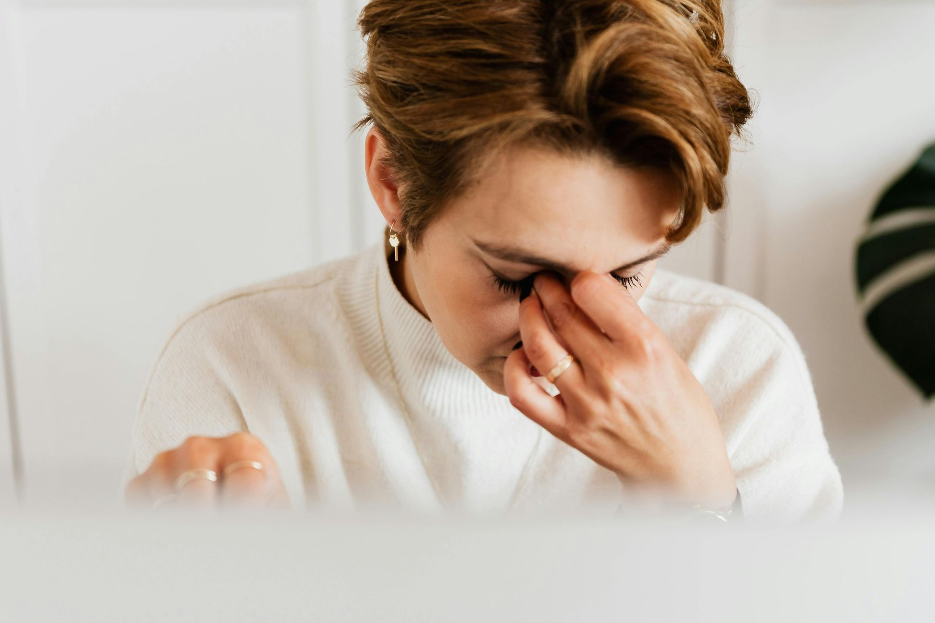Woman in a white sweater rubbing her eyes, looking tired at a desk