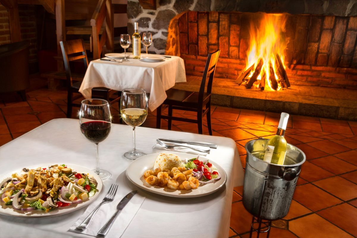 A table with plates of food and glasses of wine in front of a fireplace.