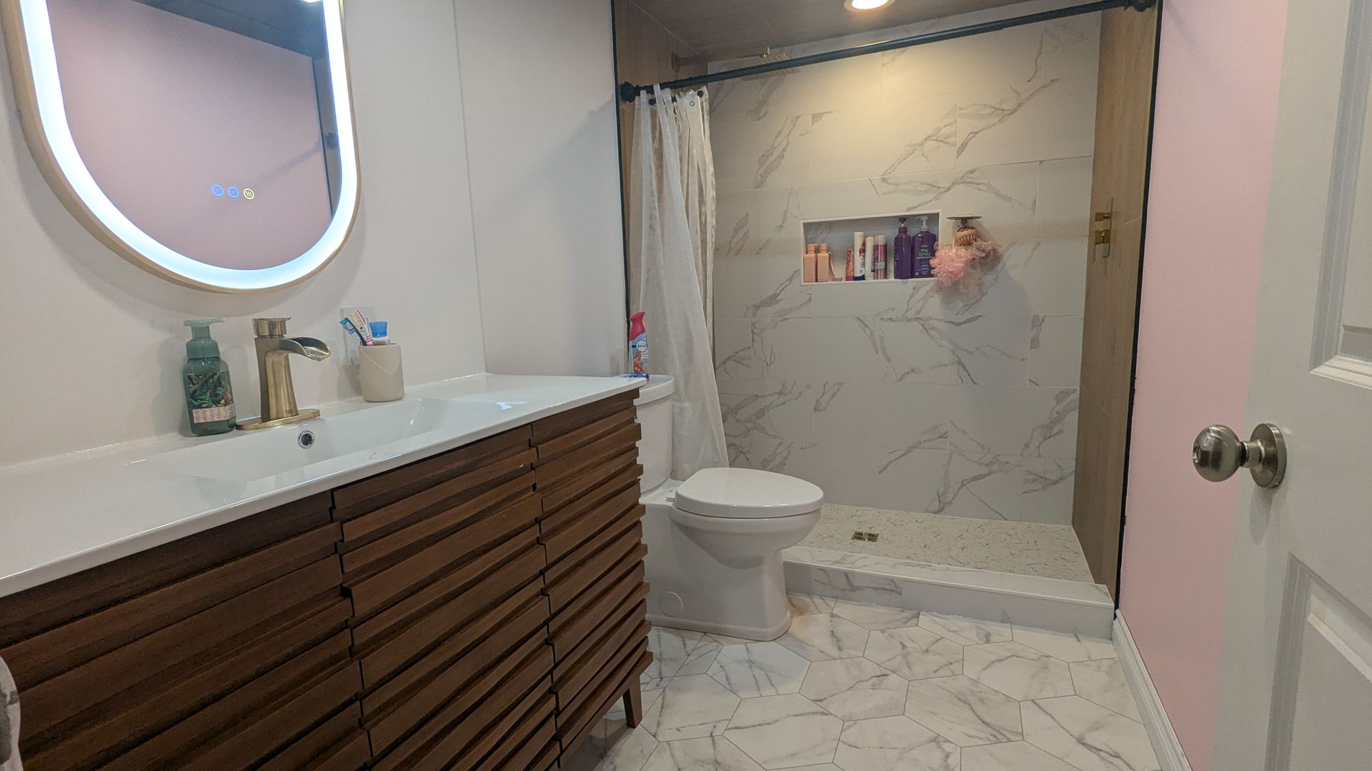 Salle de bains avec meuble-lavabo, miroir, toilettes et douche en marbre. Murs roses, robinetterie dorée et armoire en bois.