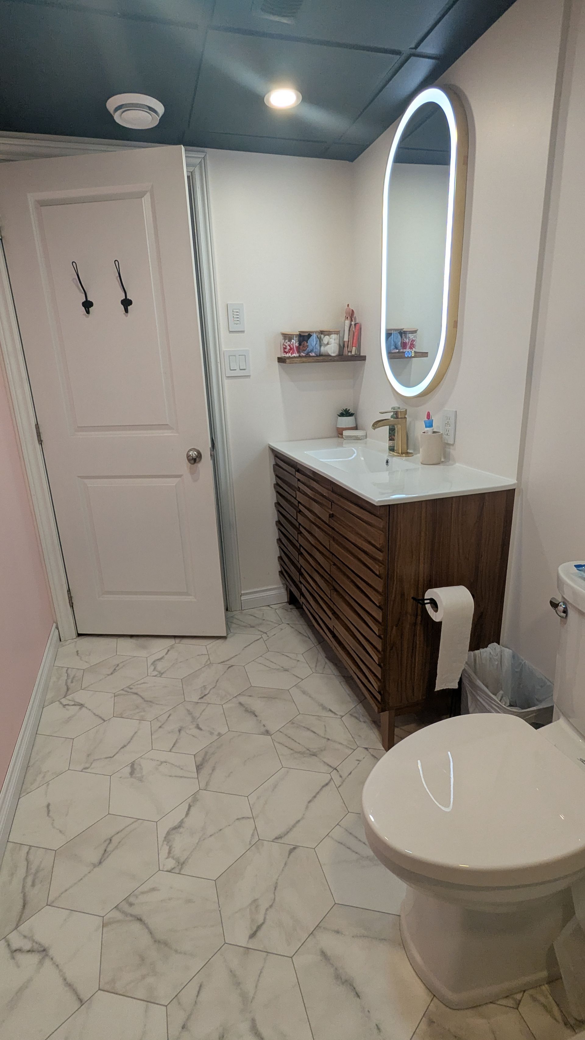 Salle de bains avec meuble-lavabo en bois, miroir ovale, toilettes blanches et sol imitation marbre.