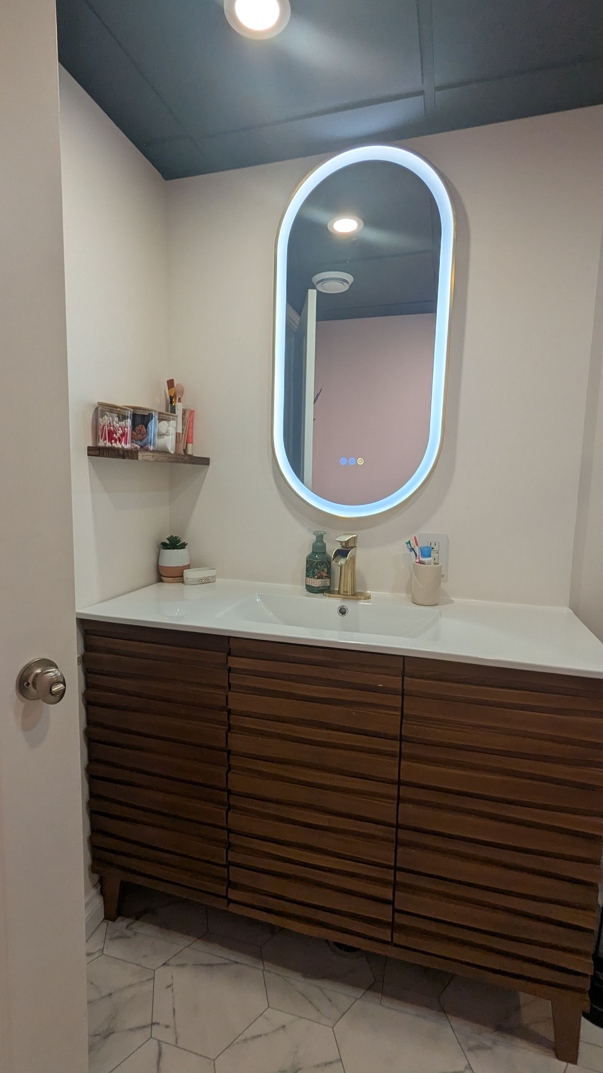 Salle de bains avec meuble-lavabo en bois, miroir ovale avec éclairage LED, murs roses et plafond foncé.