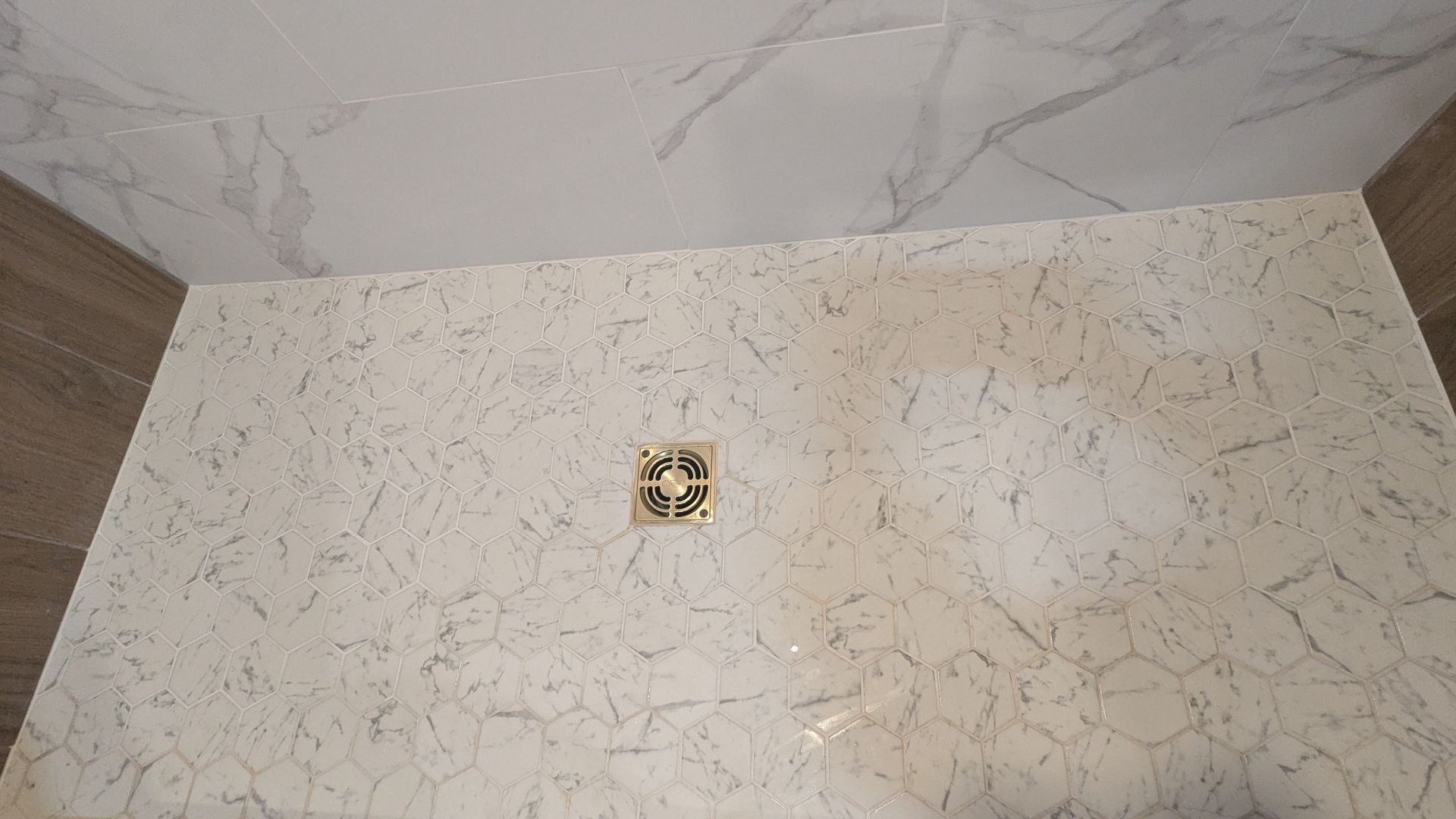 Douche carrelée en marbre blanc avec bonde dorée et éléments en bois brun.