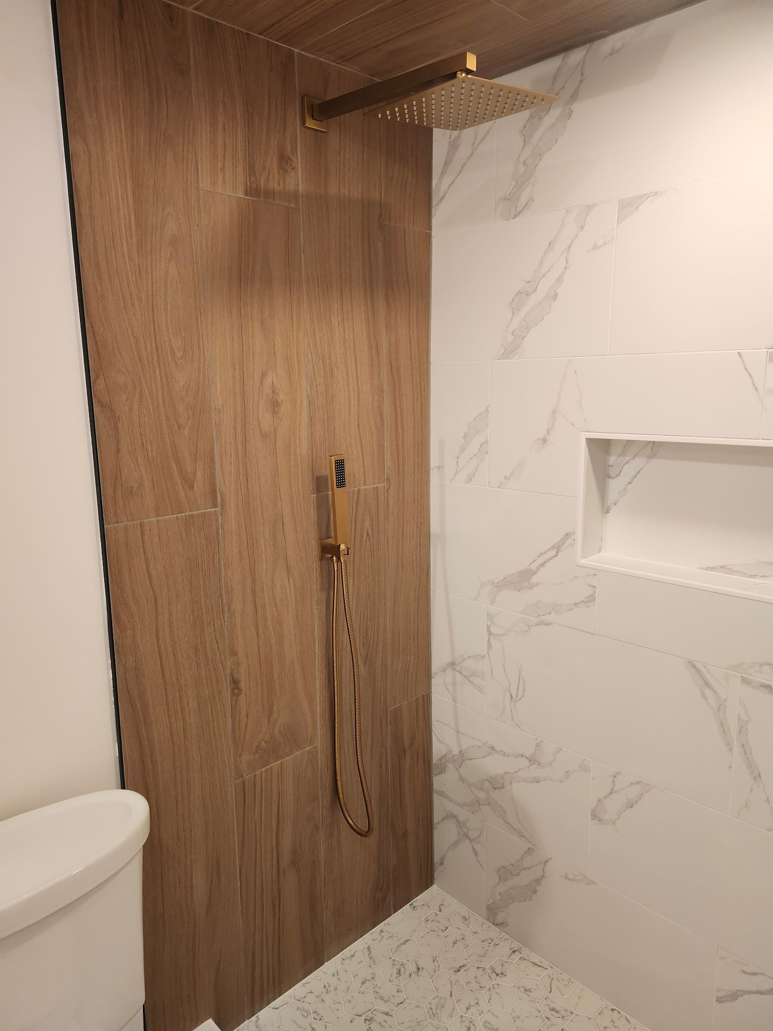Salle de bains avec mur carrelé imitation bois, robinetterie de douche dorée et mur imitation marbre blanc avec étagère.