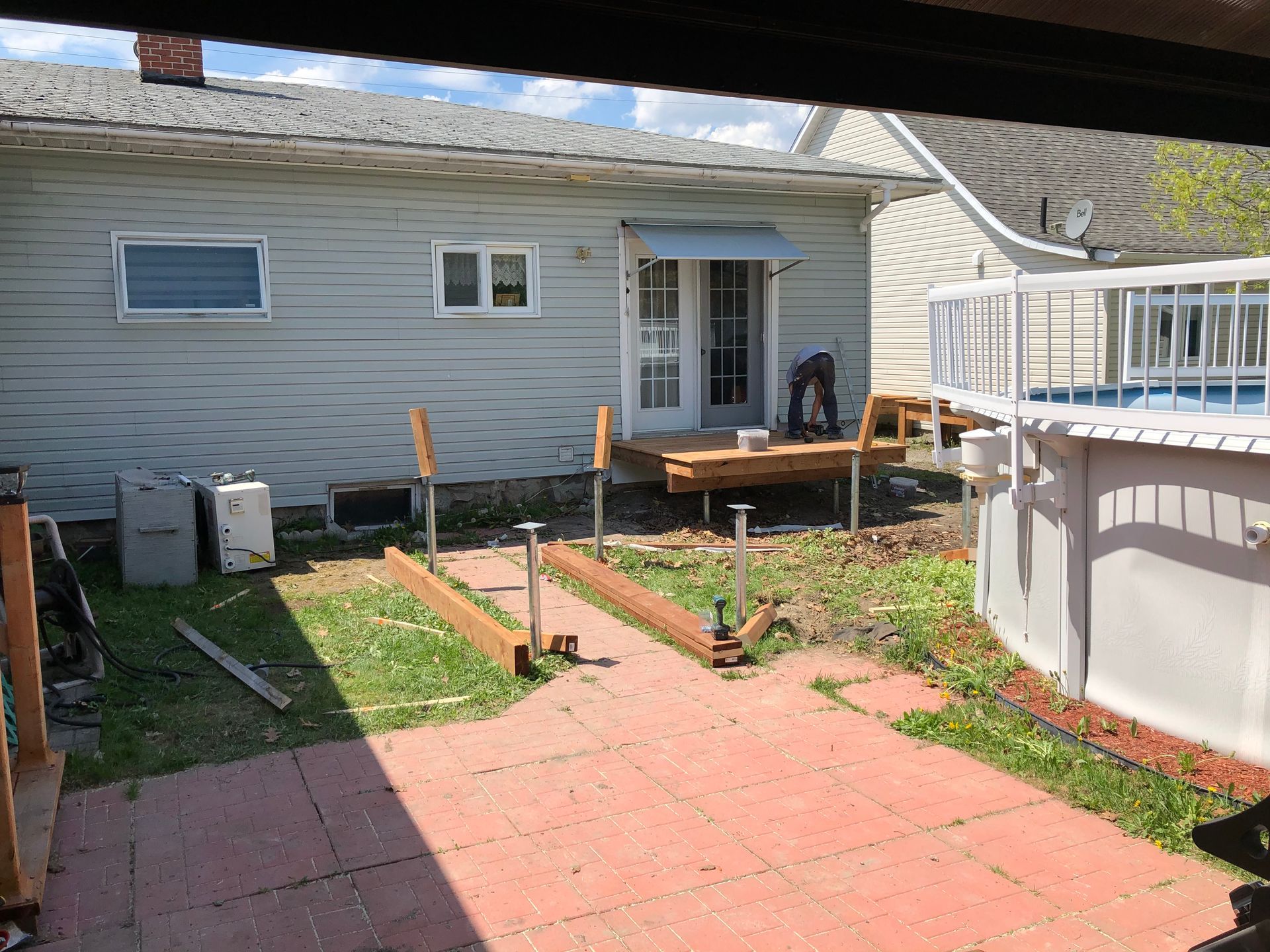 Dans un jardin, une personne travaille sur une terrasse en bois en construction, à côté d'une piscine.