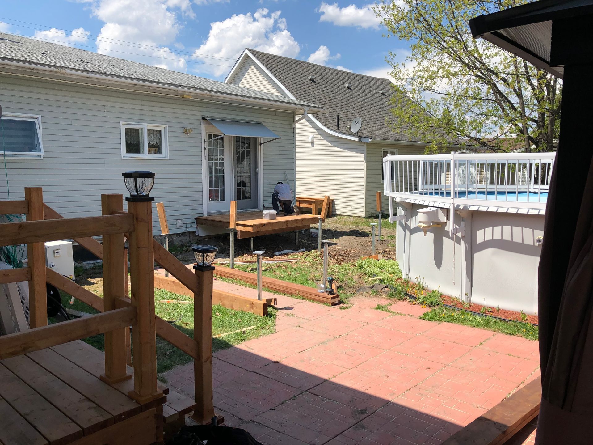 Jardin avec terrasse, piscine hors sol et maison bleu clair à deux étages sous un ciel partiellement nuageux.